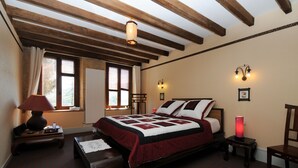 Een slaapkamer met een groot bed, houten balken in het plafond, een nachtkastje met een lamp, een stoel en een nachttafeltje.