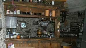 Een rustieke bar met houten planken, flessen en glazen. Er staat een ronde houten tafel met een menukaart en een kleine oranje kan.