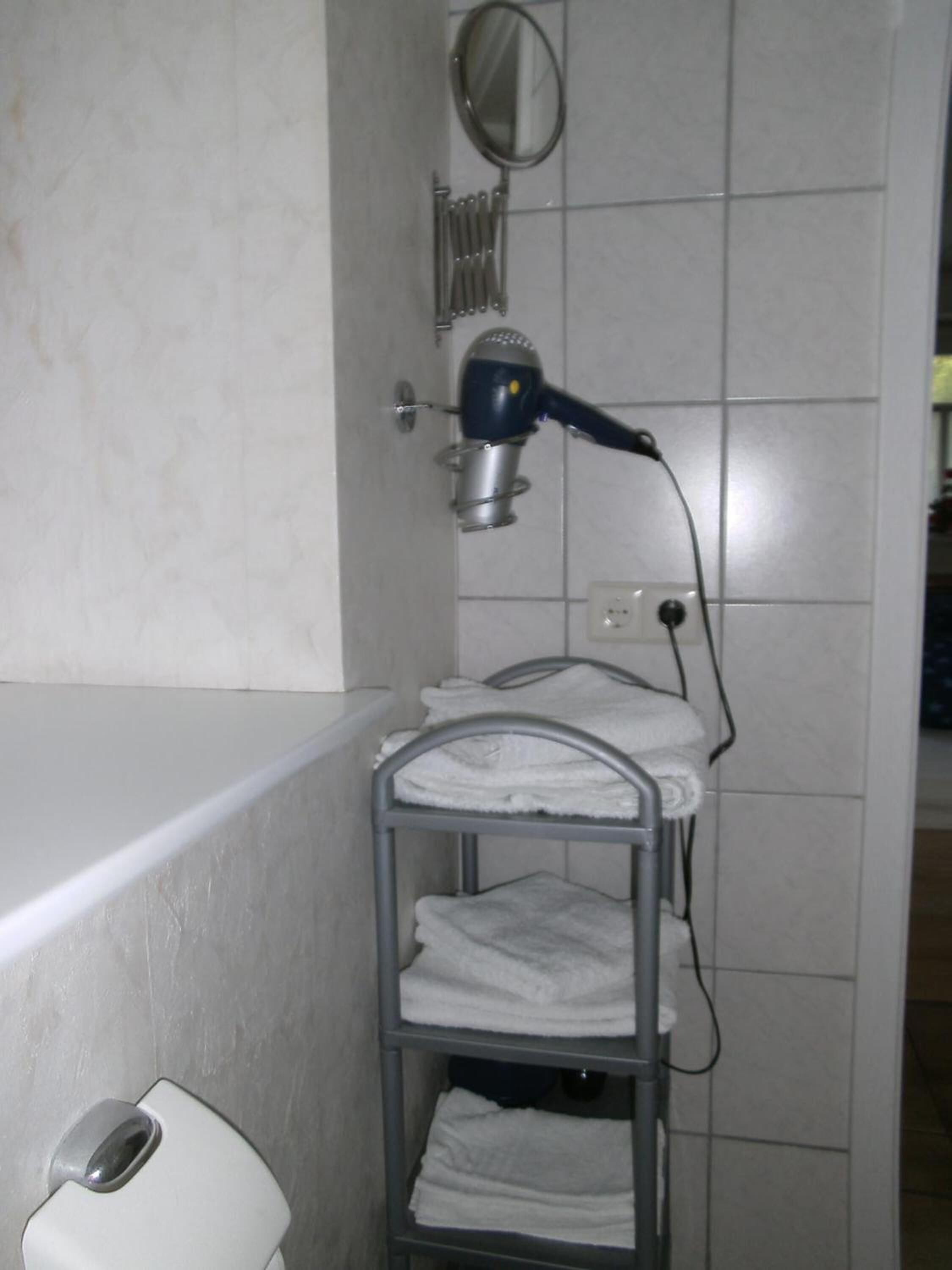 Une salle de bain équipée d’un sèche-cheveux fixé sur un support mural, d’un porte-serviettes avec des serviettes pliées, et d’un sèche-mains posé sur le sol.