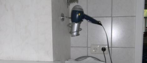 Une salle de bain équipée d’un sèche-cheveux fixé sur un support mural, d’un porte-serviettes avec des serviettes pliées, et d’un sèche-mains posé sur le sol.