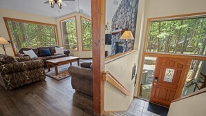 Un salon spacieux avec un grand canapé, une table basse et une cheminée. De grandes fenêtres et un escalier en bois mènent à une porte.