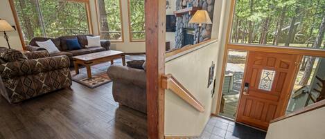 Un salon spacieux avec un grand canapé, une table basse et une cheminée. De grandes fenêtres et un escalier en bois mènent à une porte.