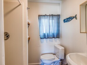 A small bathroom with a toilet, a sink, and a window with plaid curtains. - W3 - Humpback - Whale Inn (Depoe Bay)