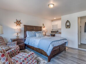 A bedroom with a wooden bed, two bedside tables with lamps, a decorative anchor wall piece, and a small kitchen area with cabinets. - W3 - Humpback - Whale Inn (Depoe Bay)
