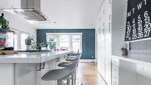A modern kitchen with white cabinetry, a central island with bar stools, and a calendar on the wall. - Sunny Family Home in Heart of Kitsilano with Mountain Views (Vancouver)