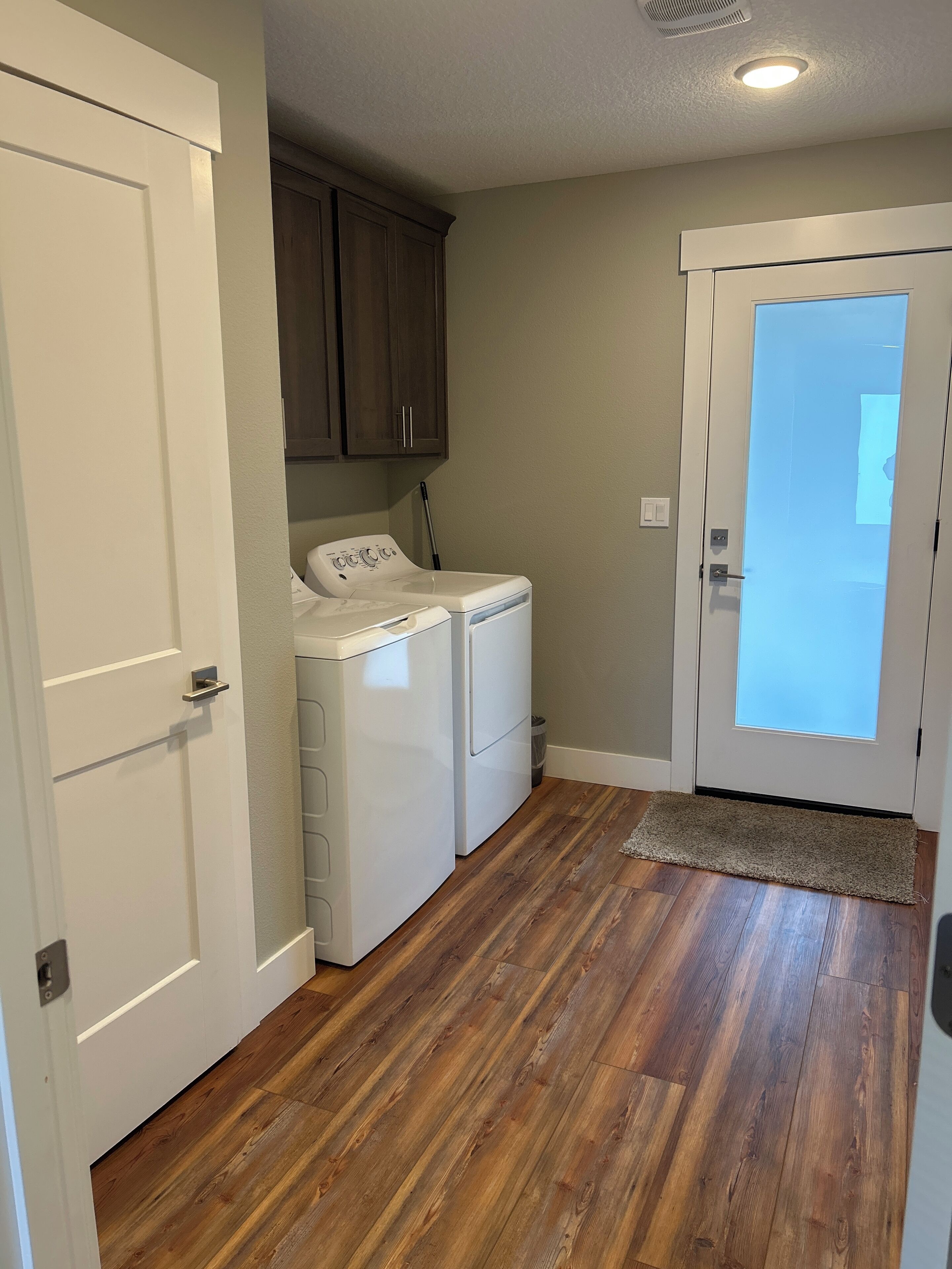A laundry room with a washing machine and dryer, wooden flooring, and a door leading to another room.