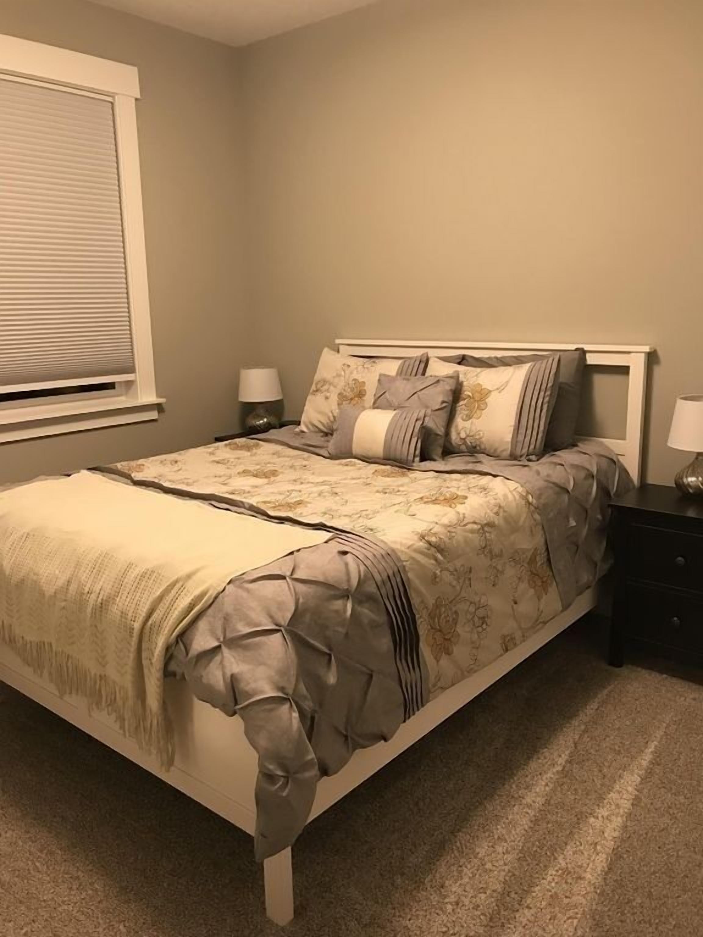 A bedroom with a bed, two bedside tables, a window with blinds, and a carpeted floor.