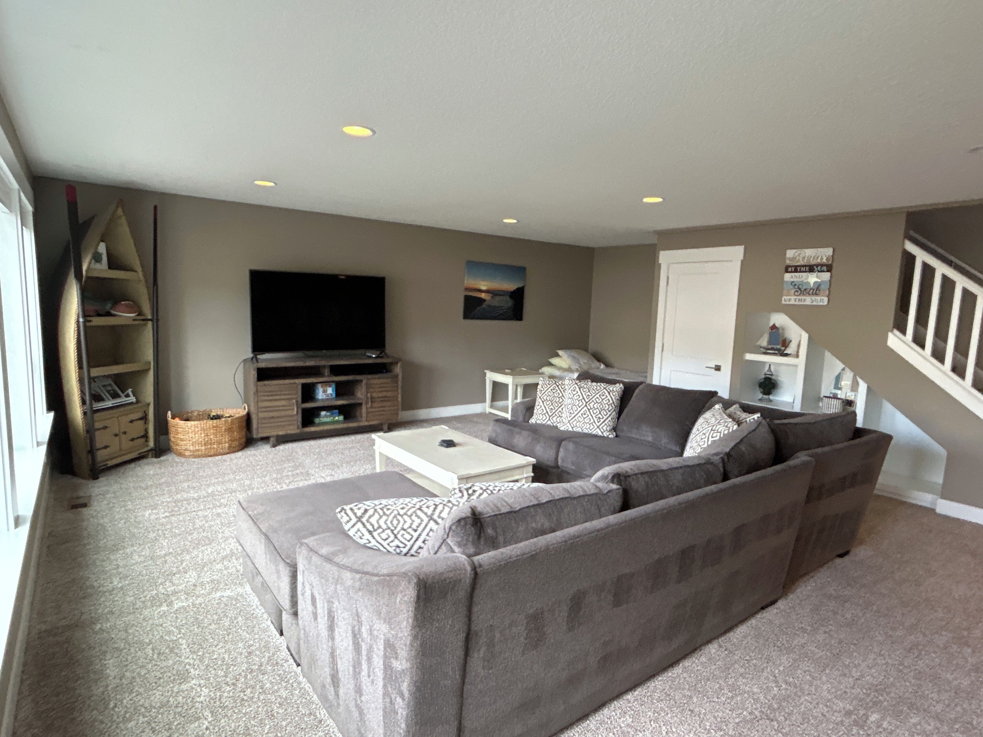 Ein Wohnzimmer mit grauem Ecksofa, einem Flachbildfernseher auf einem Holzbodenständer, einem weißen Couchtisch und einer Treppe, die zu einer oberen Etage führt.