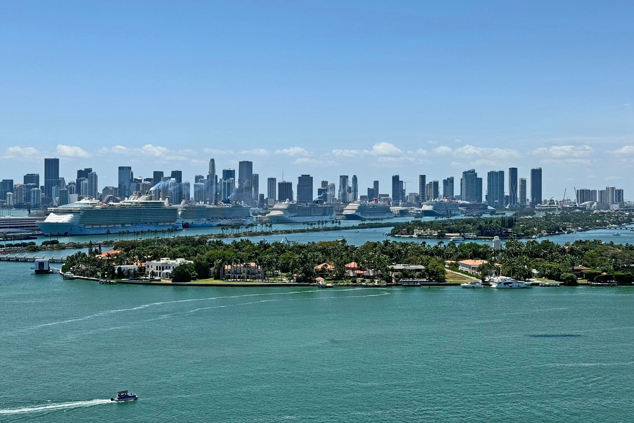 A coastal cityscape with a prominent cruise ship docked near the shore, surrounded by dense urban buildings and a small island with residential buildings.