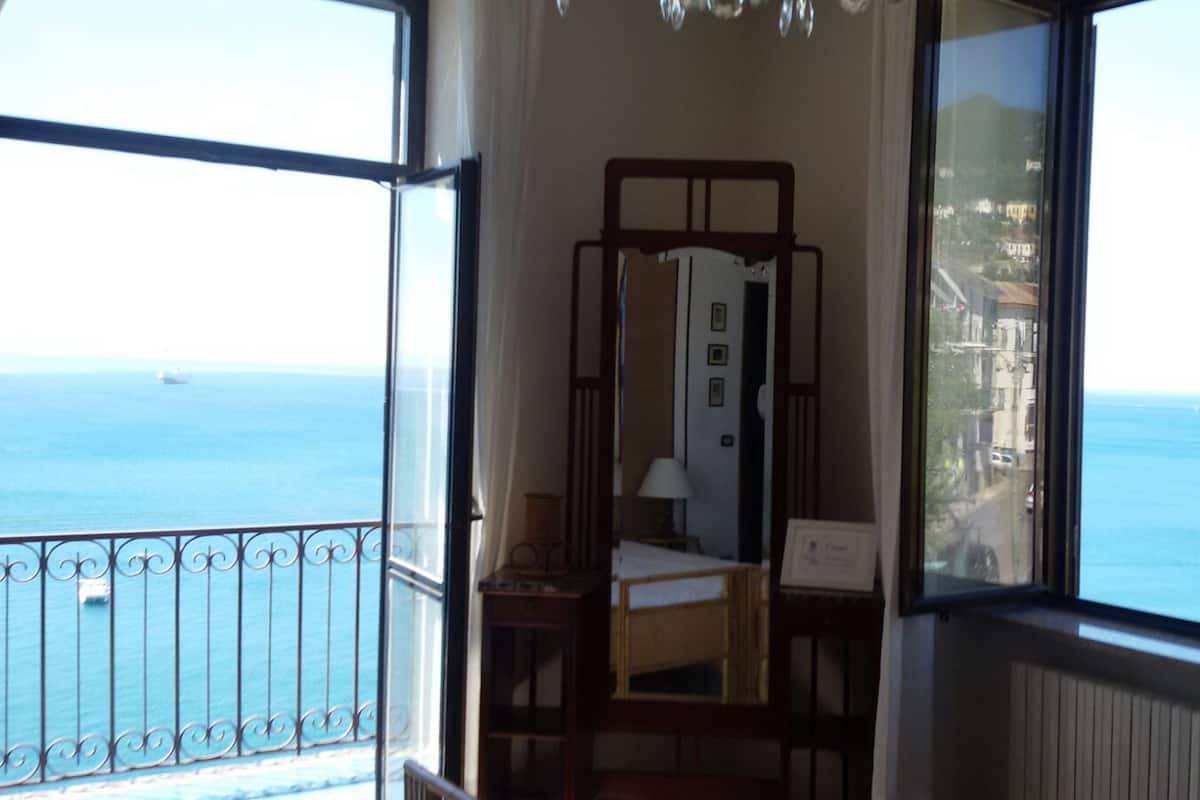 A room with a bed, a bedside table, a mirror, and a window with a view of the sea.