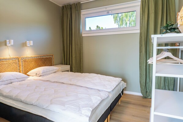 A double bed with white bedding, a bedside table, a window with curtains, and a white wardrobe with hanging space.