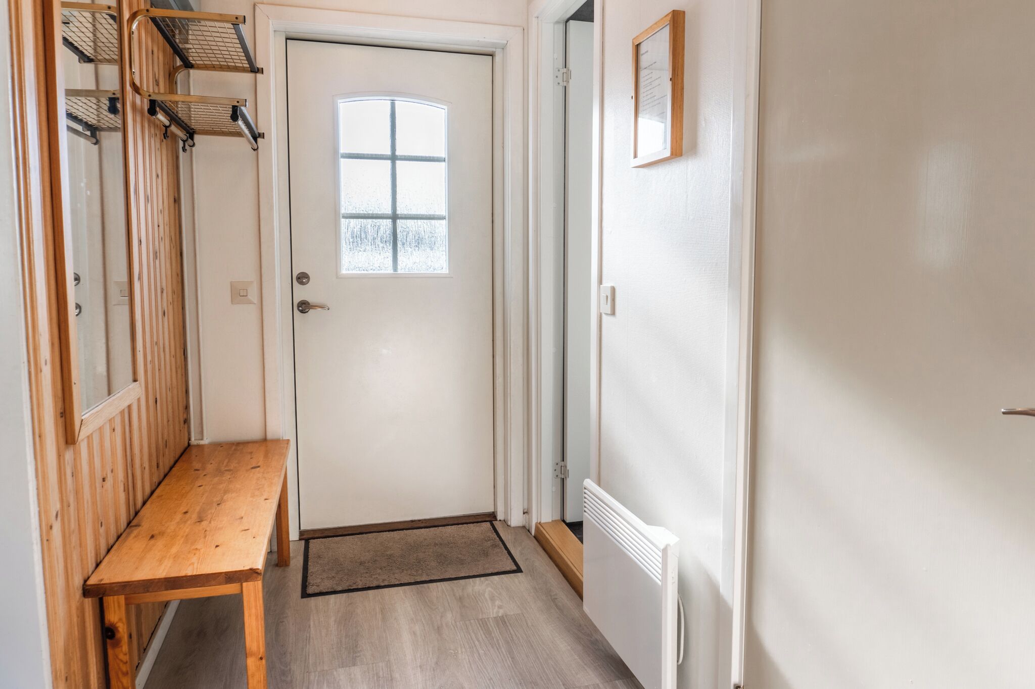 A narrow hallway with a wooden bench, a white door with a window, and a radiator on the wall.