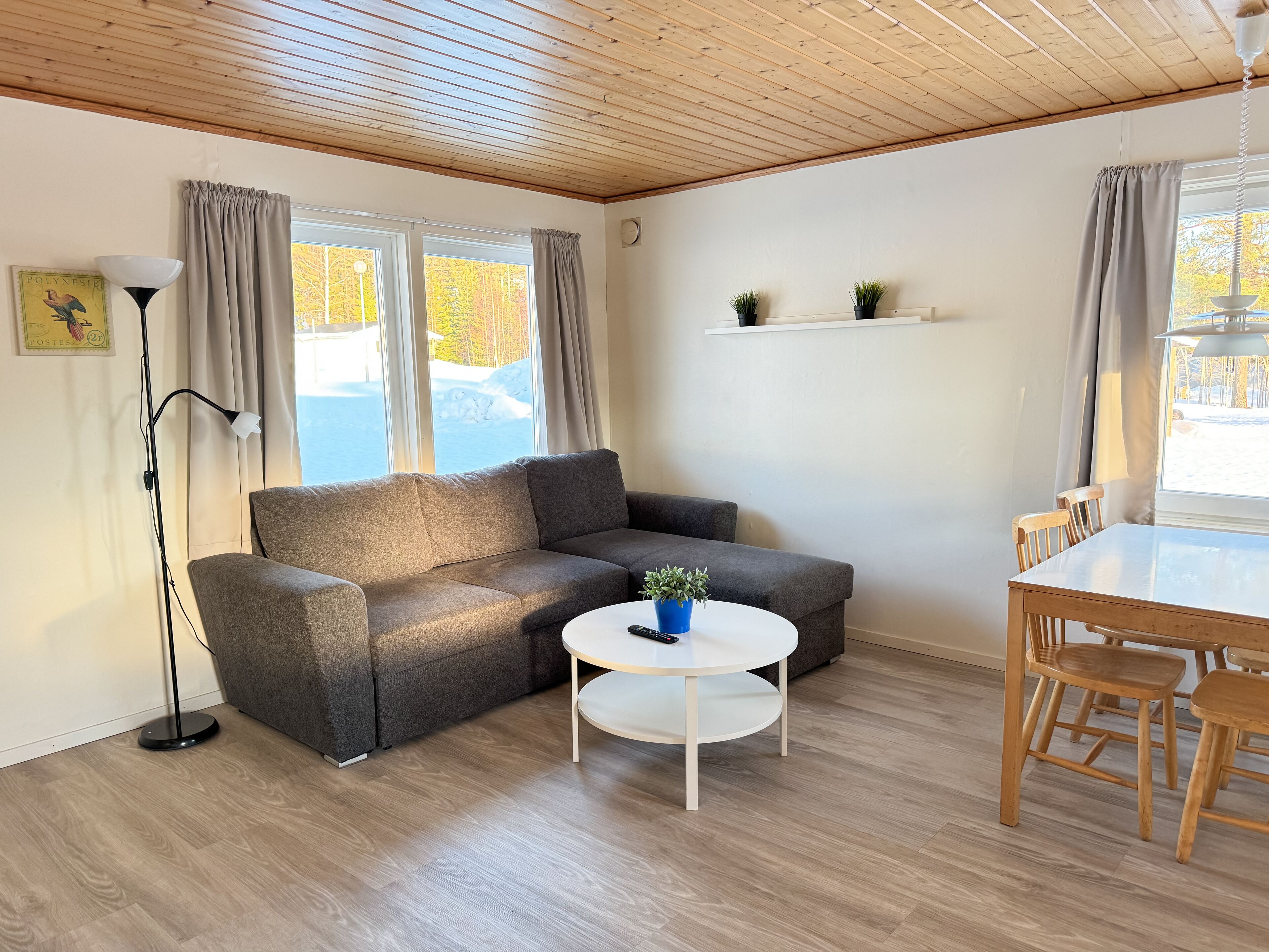 Un salon chaleureux avec un canapé gris, une table basse ronde et une table à manger avec des chaises. Il y a deux fenêtres avec des rideaux, une lampe sur pied et une petite étagère avec des plantes.