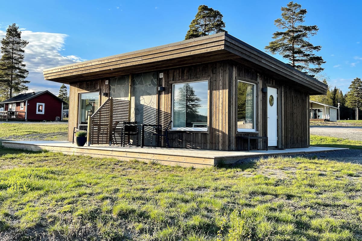 Eine moderne Holzblockhütte mit großen Fenstern, einer Veranda mit Stühlen und einem roten Haus im Hintergrund.