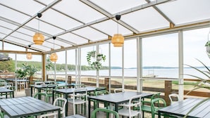 A covered outdoor dining area with tables and chairs, large windows, and hanging plants. - Gullviks Havsbad (Domsjö)