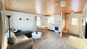 Una sala de estar acogedora con un sofá, una mesa de centro y un comedor con sillas. Se ve una cocina con alacenas y electrodomésticos blancos.