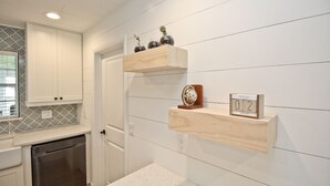 A kitchen with white cabinets, a stainless steel dishwasher, wooden shelves with decorative items, and a wall-mounted clock. - Sunshine Stay, Central Fort Myers Gated Yard (Fort Myers)