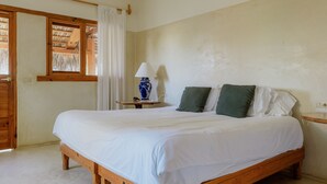 A wooden bed with white bedding, a nightstand with a lamp, and a window with curtains. - Villa Gardenia - Oceanfront House Mango (Todos Santos)