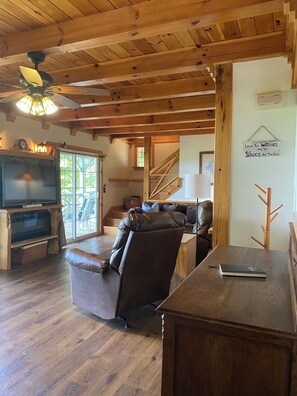 A cozy living room with a wooden ceiling, a leather sofa, a TV, and a staircase leading upstairs.