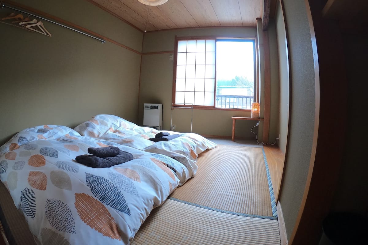 A room with two beds, a window, a heater, and a small table with a lamp.