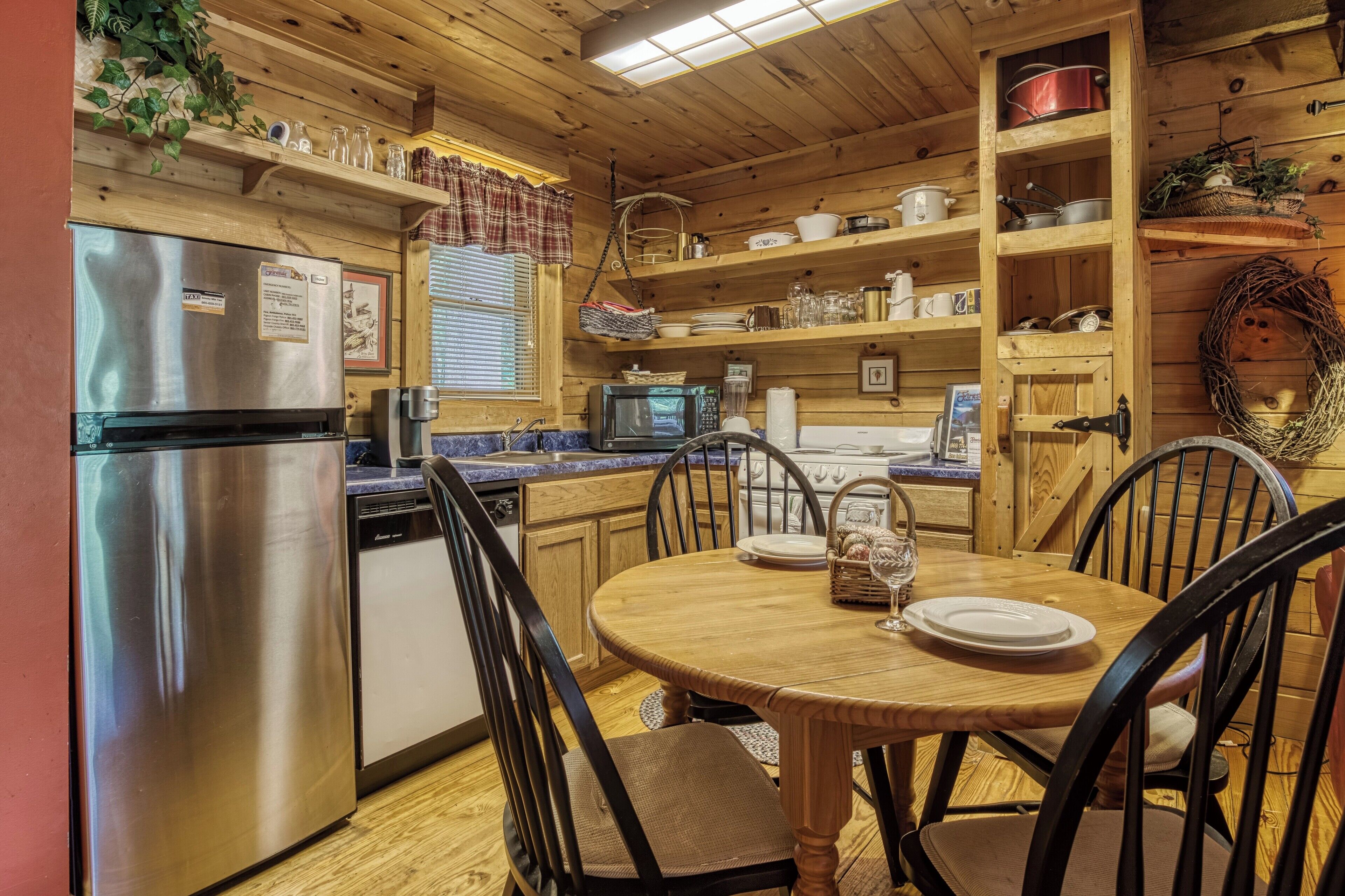 Cucina di una baita in legno con frigorifero in acciaio inossidabile, forno a microonde e lavastoviglie. Si nota un tavolo rotondo in legno con sedie, e ripiani sopra il bancone con piatti e pentole.