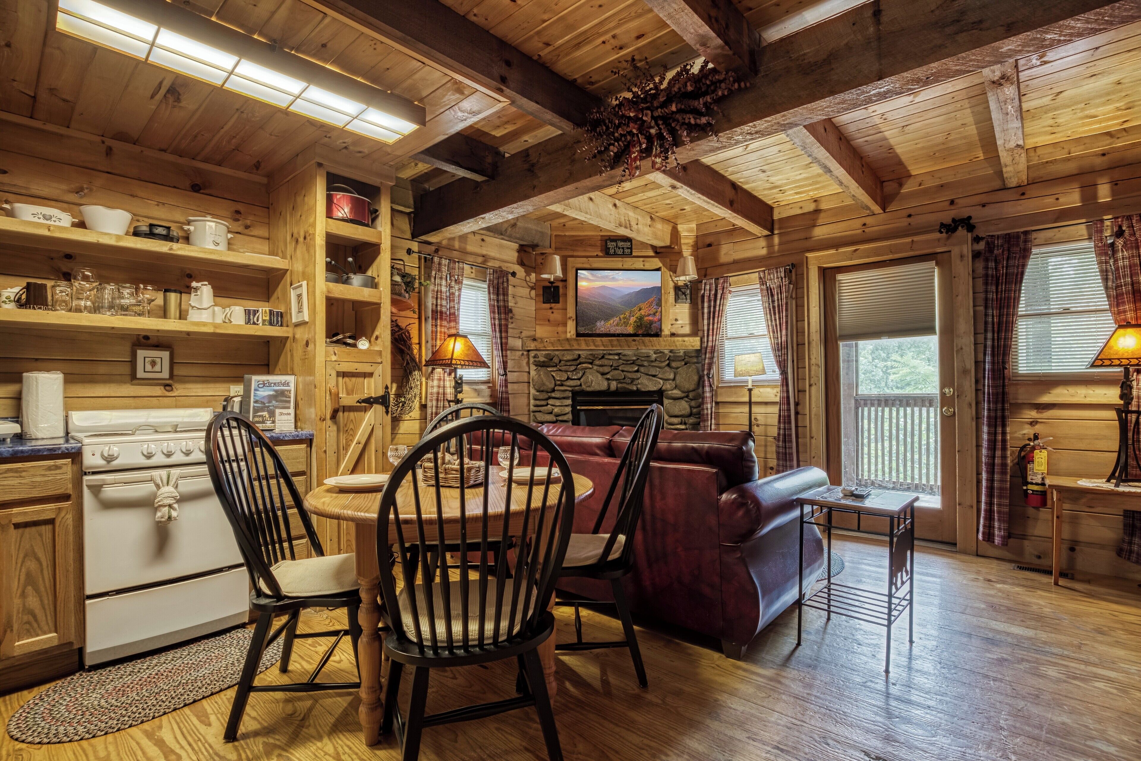 A rustic cabin interior with wooden walls, a stone fireplace, a dining area with a table and chairs, a red leather sofa, and a kitchen with a stove and shelves.
