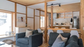 Uma sala de estar moderna com sofás cinzas, uma mesa de centro de vidro e uma área de jantar de madeira, com uma cozinha ao fundo.