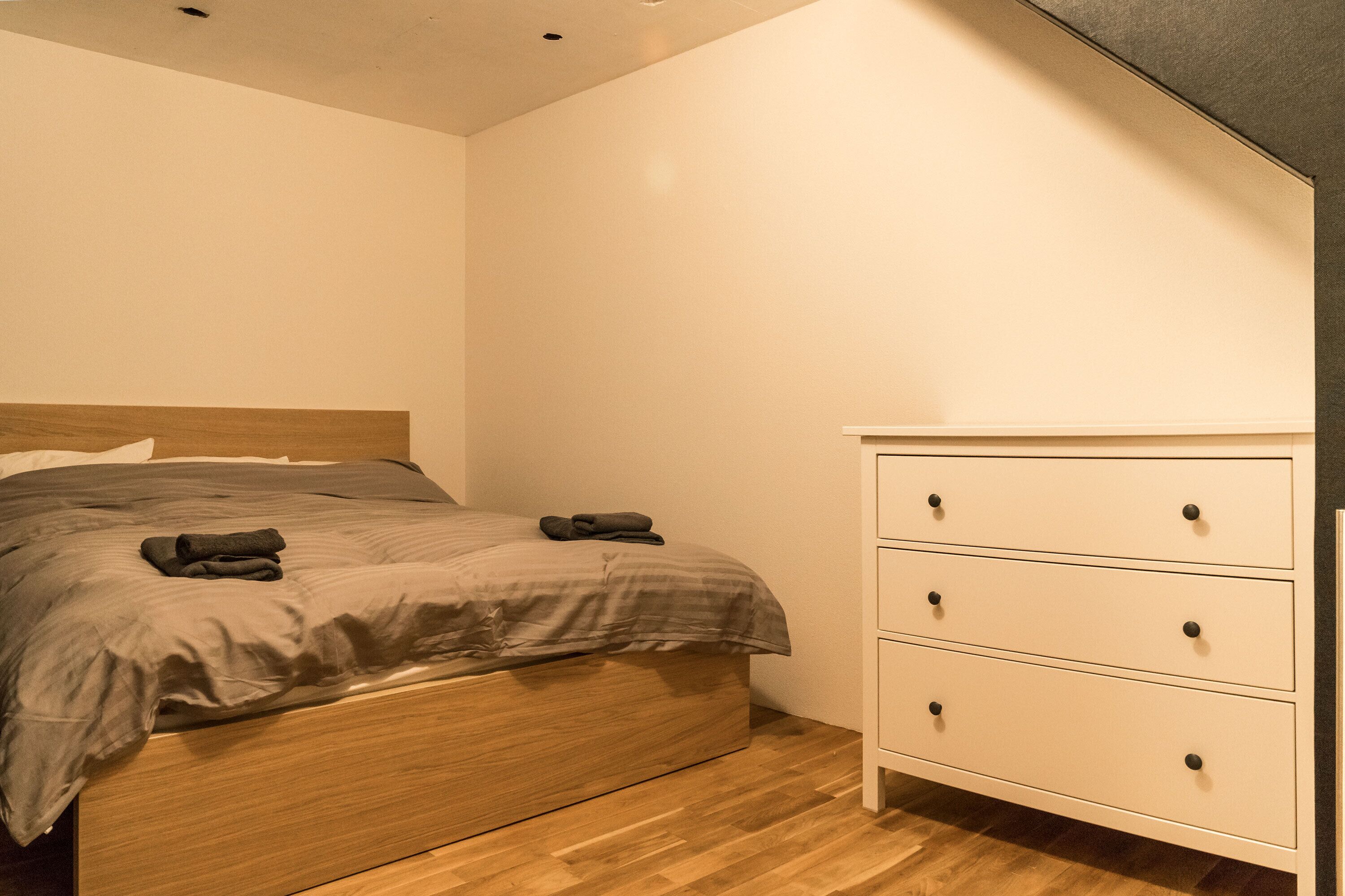 A bedroom with a wooden bed, a white dresser with drawers, and a wooden floor.