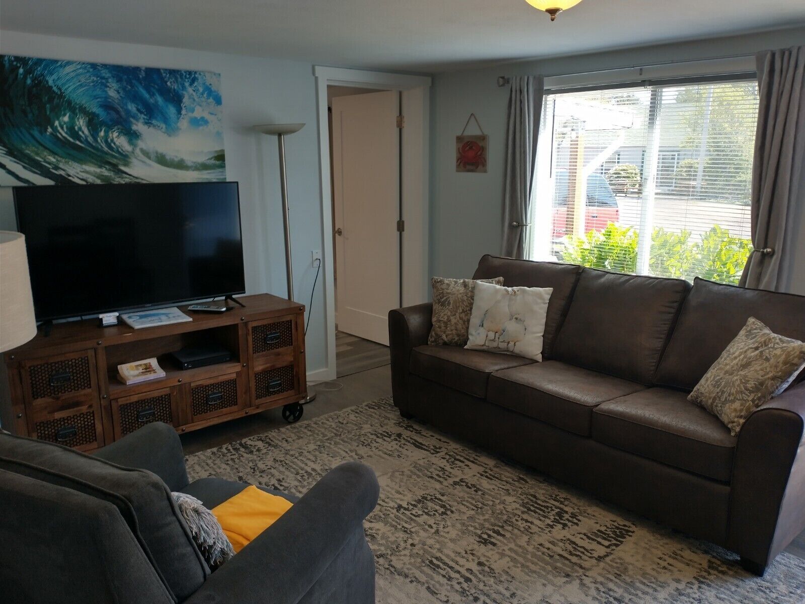 A living room with a brown leather sofa, a television on a wooden stand, a patterned rug, and a large window with curtains.