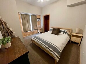 A bedroom with a bed, bedside tables, a nightstand, a window with curtains, a door, and a potted plant.