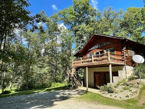 A wooden cabin with a balcony, surrounded by trees and a gravel path.