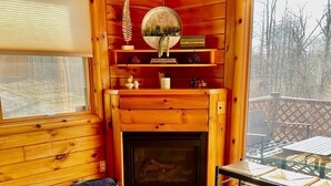 A cozy cabin interior with a fireplace, two gray armchairs, a round wooden table, and a large window overlooking a deck. - Pet friendly, hot tub, close to Hocking Hills! (McArthur)