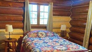 A log cabin room with a wooden bed, two bedside lamps, a window with curtains, and a wooden floor. - Private Lake Superior Log Cabin on the Beach (Au Train)