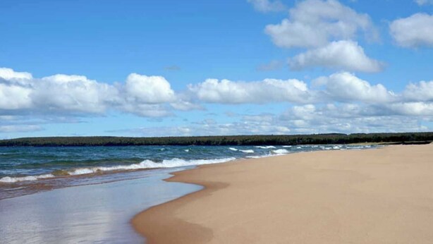 A beach with a wide sandy shore, a calm sea, and a distant forest line under a partly cloudy sky. - Private Lake Superior Log Cabin on the Beach (Au Train)