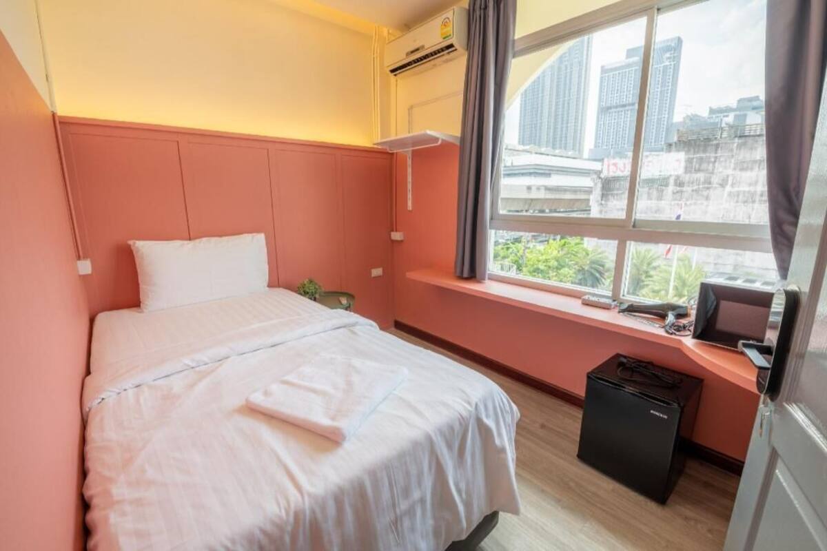 A hotel room with a bed, a window with a city view, a desk with a computer, and an air conditioning unit.