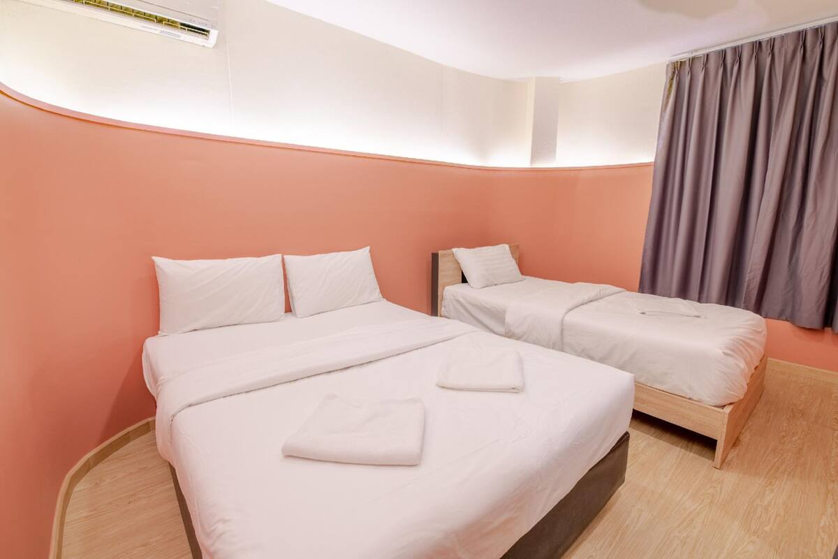 A hotel room with two beds, a wooden floor, a large window with curtains, and an air conditioning unit on the wall.