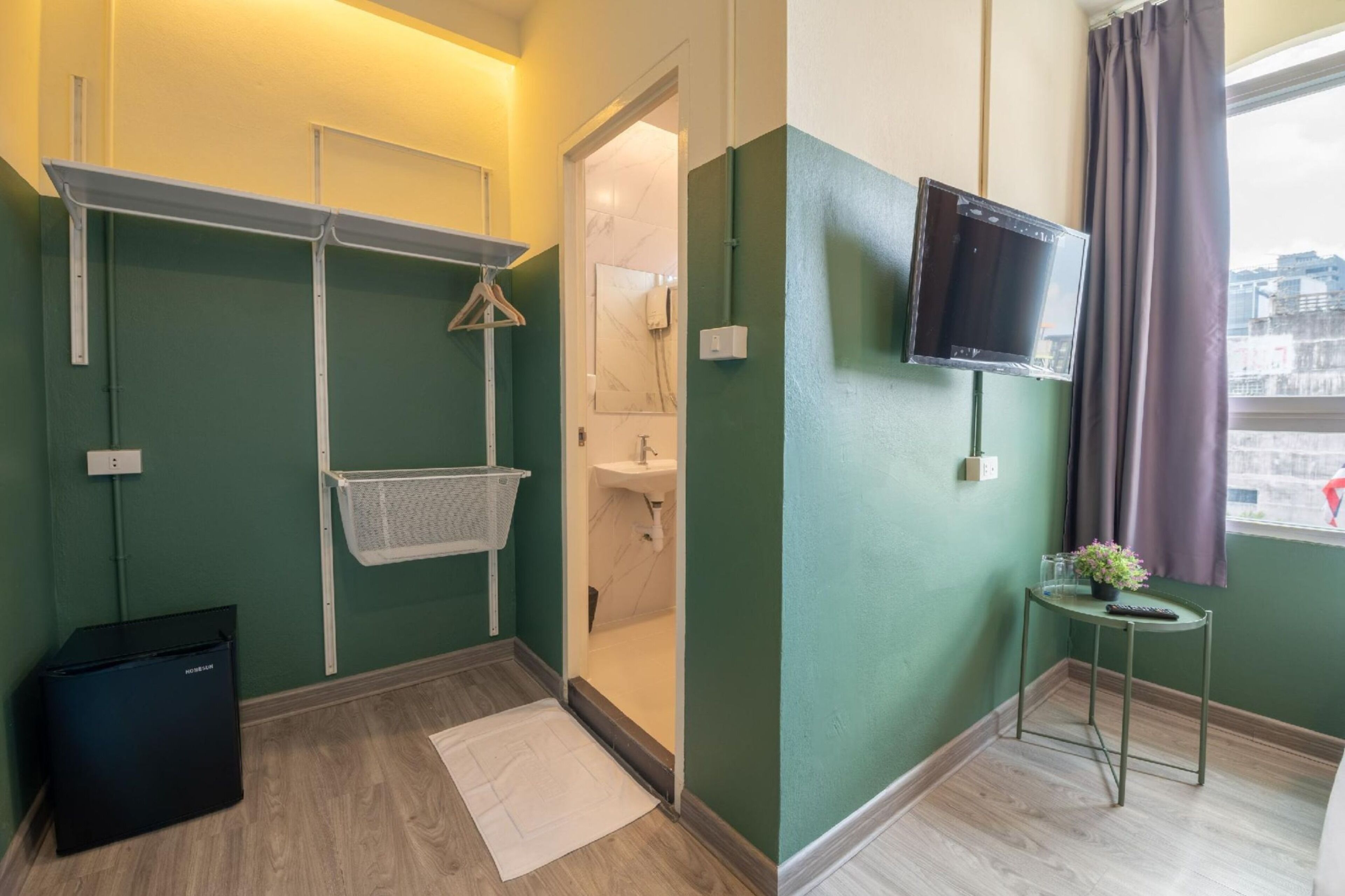 A compact hotel room with a green wall, a wooden floor, a small table with a plant, a TV mounted on the wall, and a door leading to a bathroom.