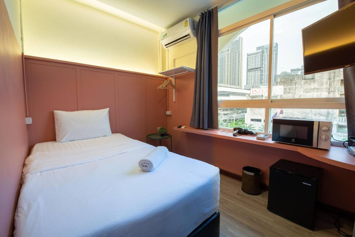 A hotel room with a bed, a small table, a microwave, a trash bin, and a window with a city view.
