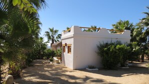 A small, white building with a thatched roof, surrounded by palm trees and a sandy ground. - Villa Gardenia - Casa Jazmín directly on the sea (Todos Santos)