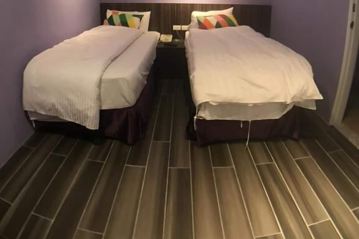A room with two beds, each with a headboard and pillows, and a nightstand between them. The room has purple walls and a wooden floor.