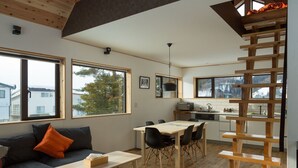 A modern interior with a sofa, dining table, and chairs, large windows, and a wooden staircase. - Harvest Eco House (Nozawaonsen)