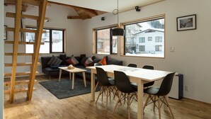 A modern living space with a wooden ladder, a sofa with cushions, a dining table with chairs, and large windows offering a view of the outside. - Harvest Eco House (Nozawaonsen)