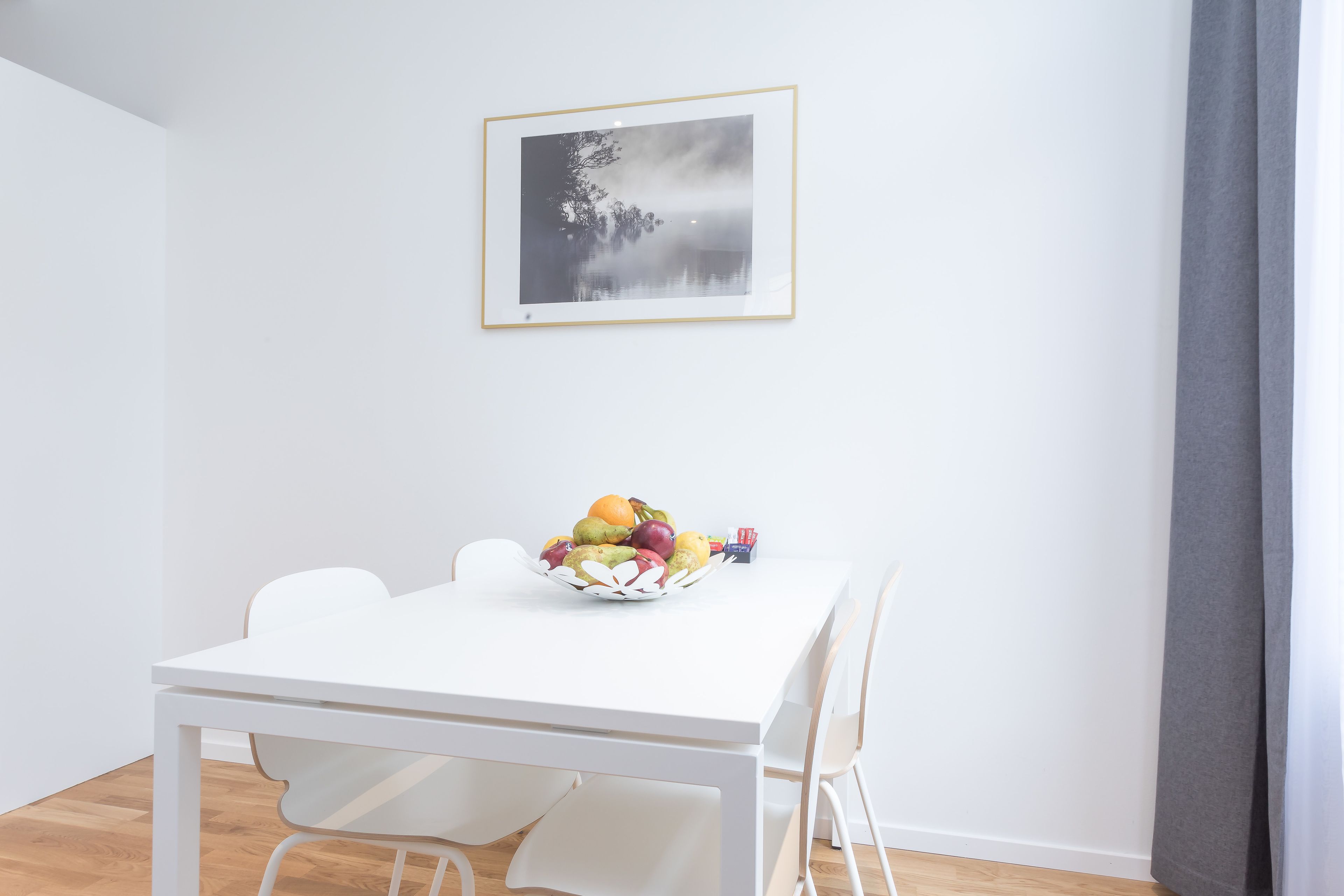 A white dining table with a fruit bowl, white chairs, and a framed picture on the wall.
