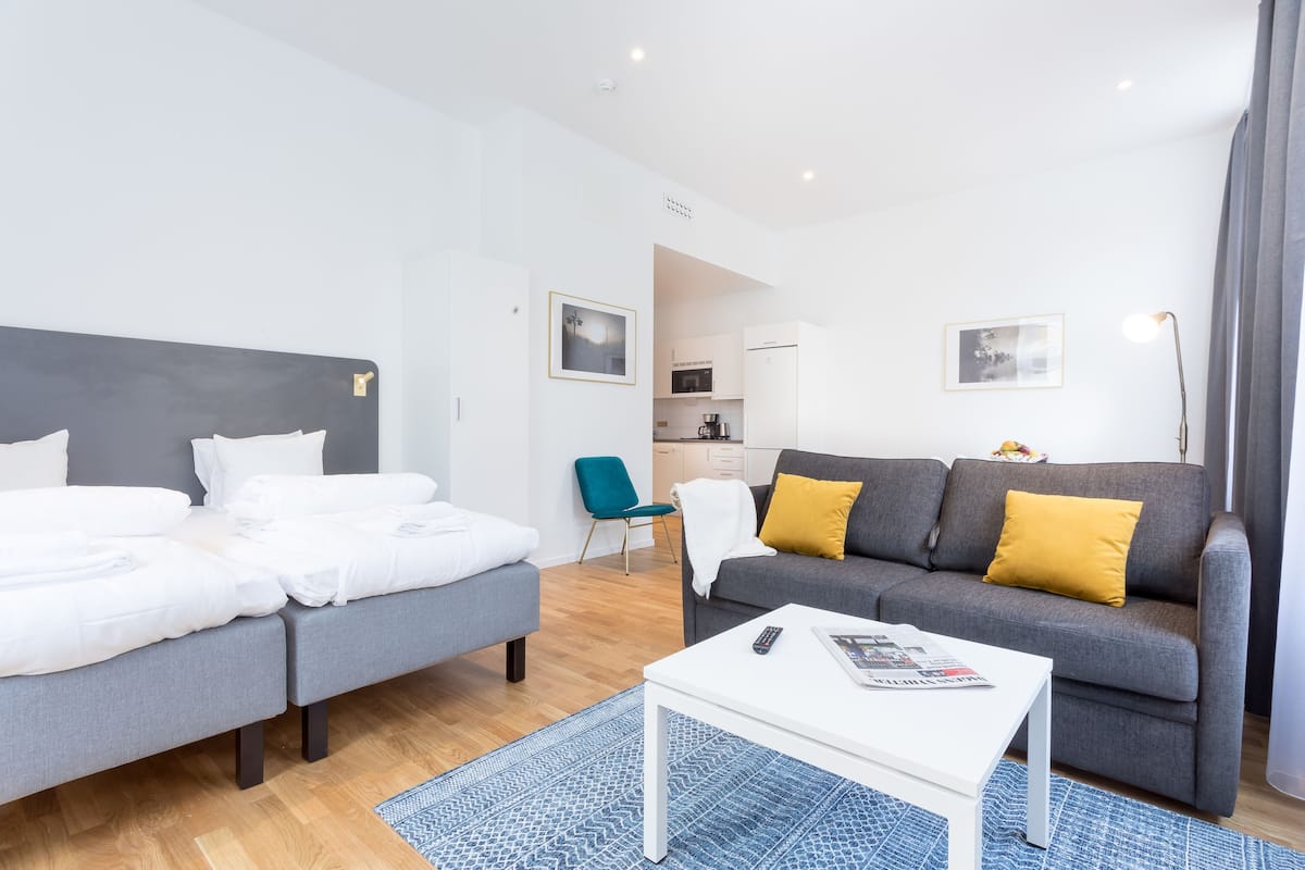 A modern hotel room with a bed, a sofa, a coffee table, and a view of the kitchen area.