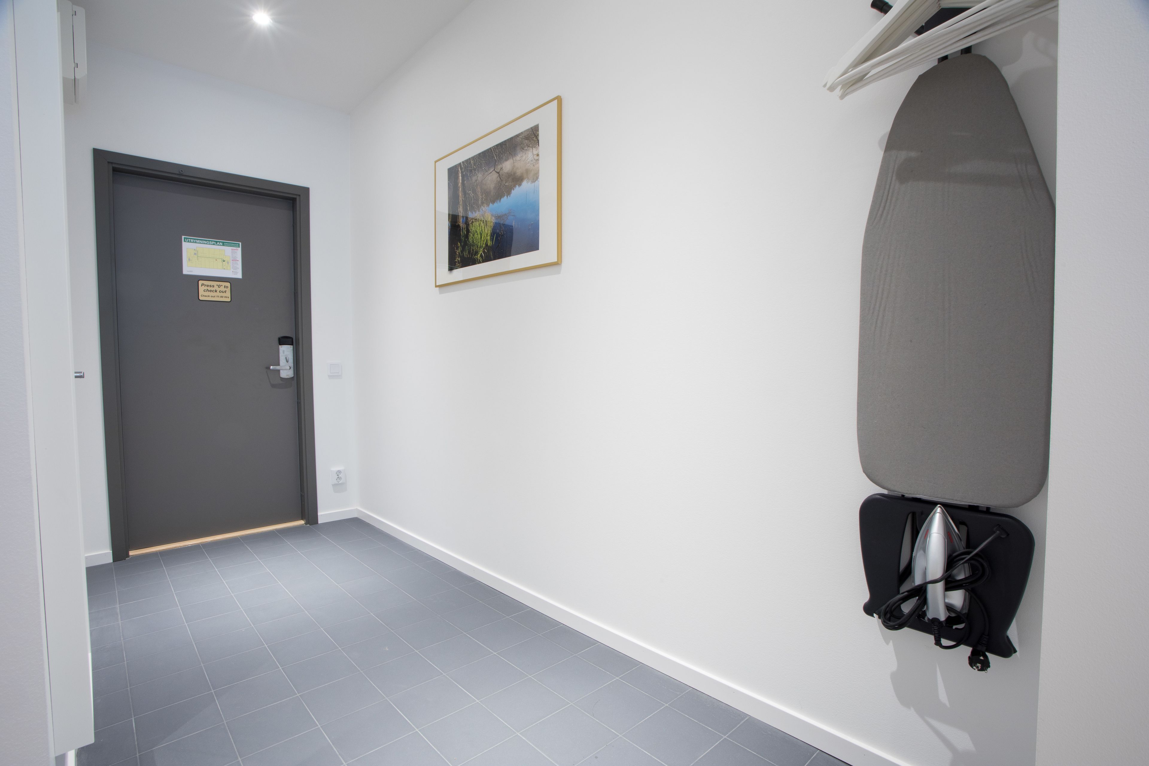 Un couloir avec une porte grise, un tableau encadré accroché au mur et une planche à repasser fixée au mur.