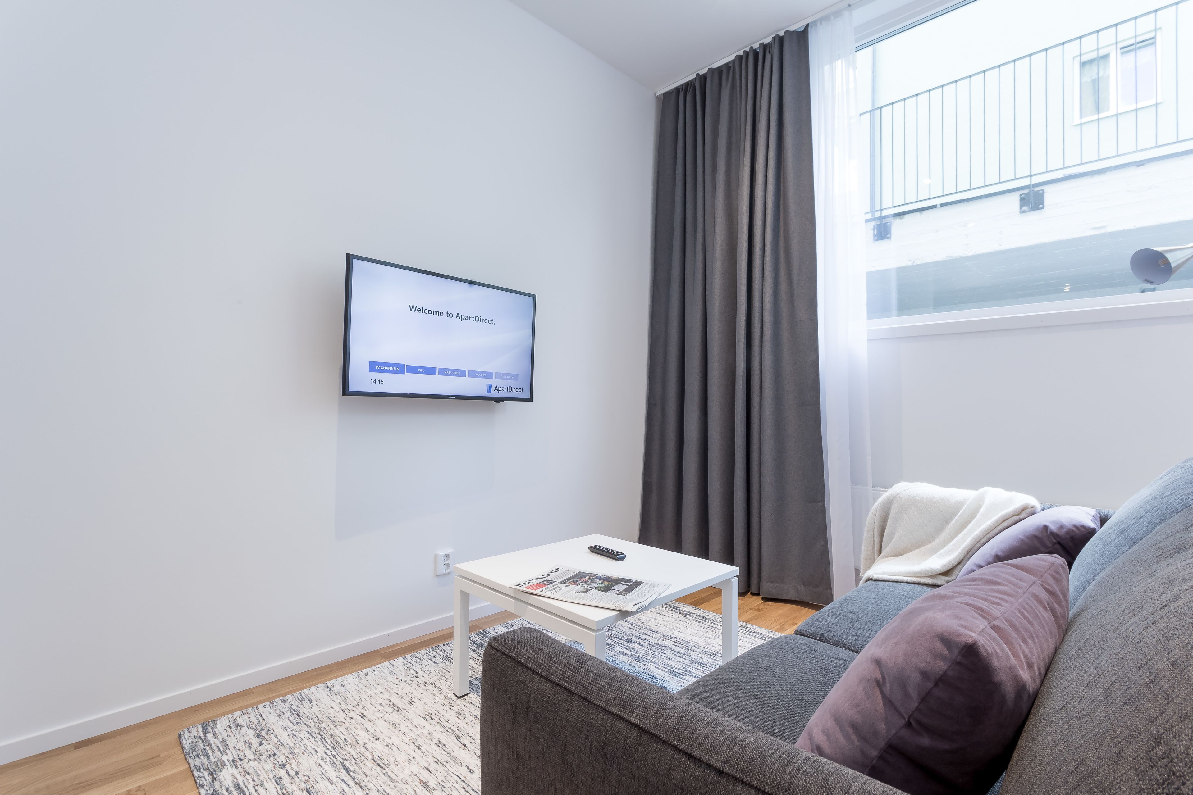 Un salon moderne avec un téléviseur à écran plat fixé au mur, une table basse blanche avec une télécommande, un canapé gris avec des coussins et une fenêtre ornée de rideaux gris.