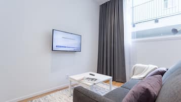 Un salon moderne avec un téléviseur à écran plat fixé au mur, une table basse blanche avec une télécommande, un canapé gris avec des coussins et une fenêtre ornée de rideaux gris.