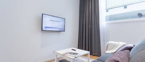 Un salon moderne avec un téléviseur à écran plat fixé au mur, une table basse blanche avec une télécommande, un canapé gris avec des coussins et une fenêtre ornée de rideaux gris.