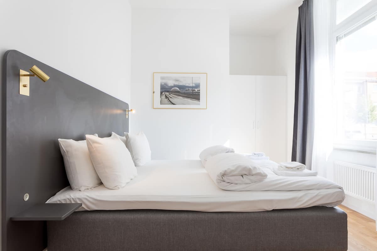 A modern hotel room with a large bed, a bedside table, a framed picture on the wall, and a view of the outdoors through a window.