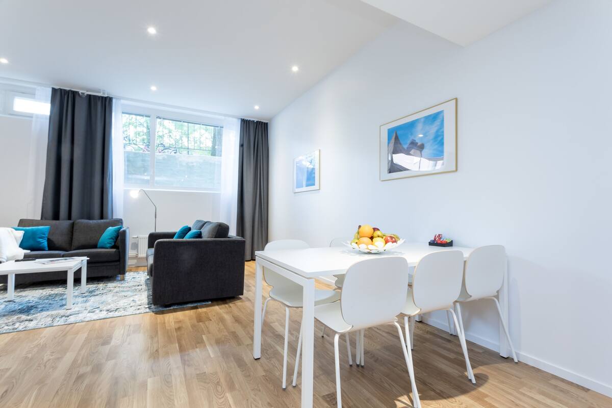 A modern living room with a white dining table, grey sofas, and a large window with curtains.